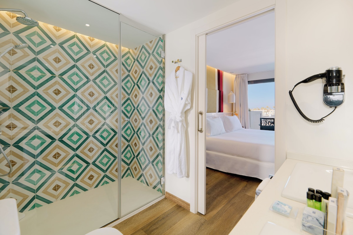 A hotel room with a patterned shower wall, a bed with white linens, a robe hanging on the door, and a hairdryer on the wall.