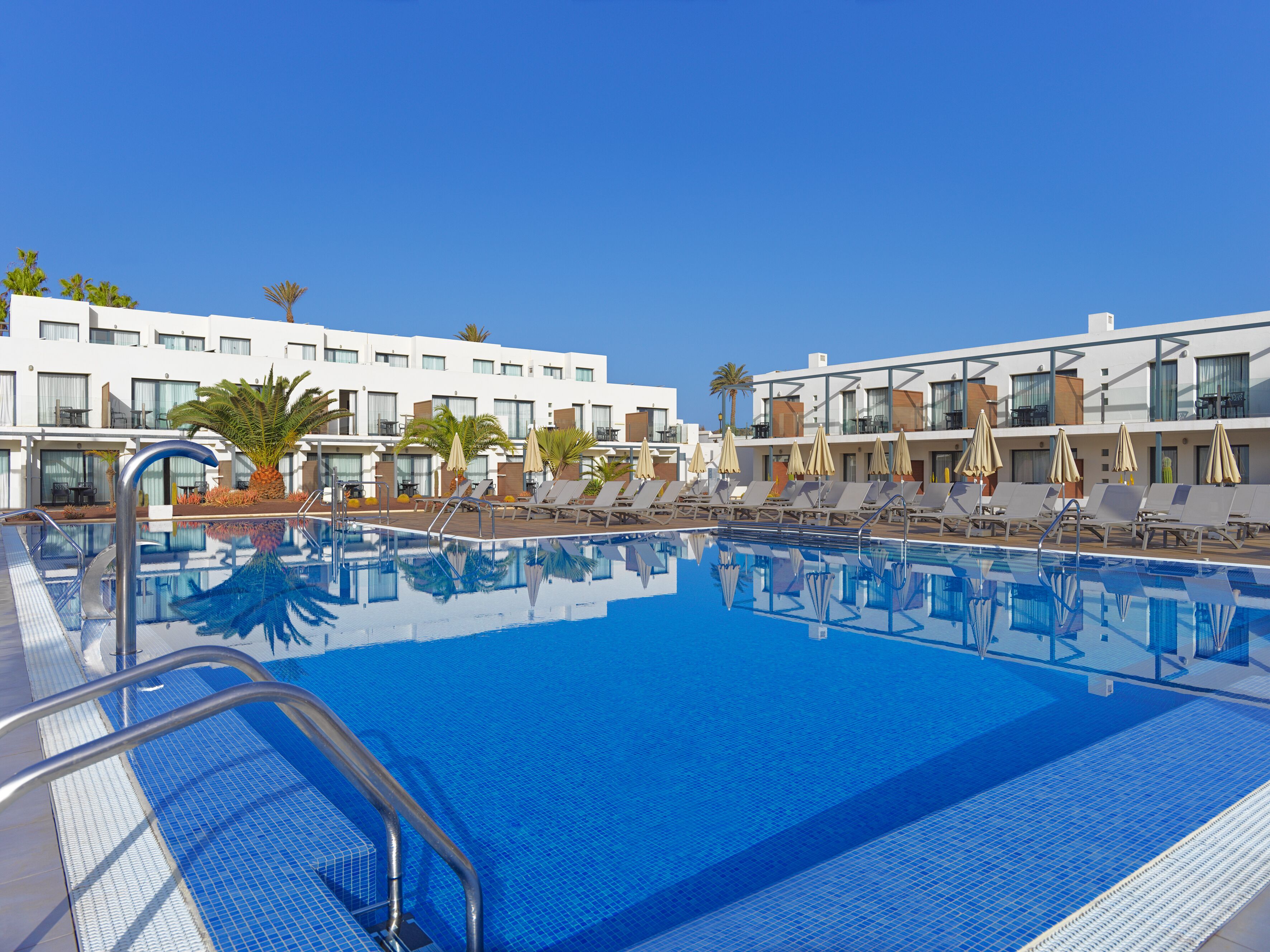 Un espace piscine d’hôtel avec une eau d’un bleu limpide, des chaises longues et des parasols.