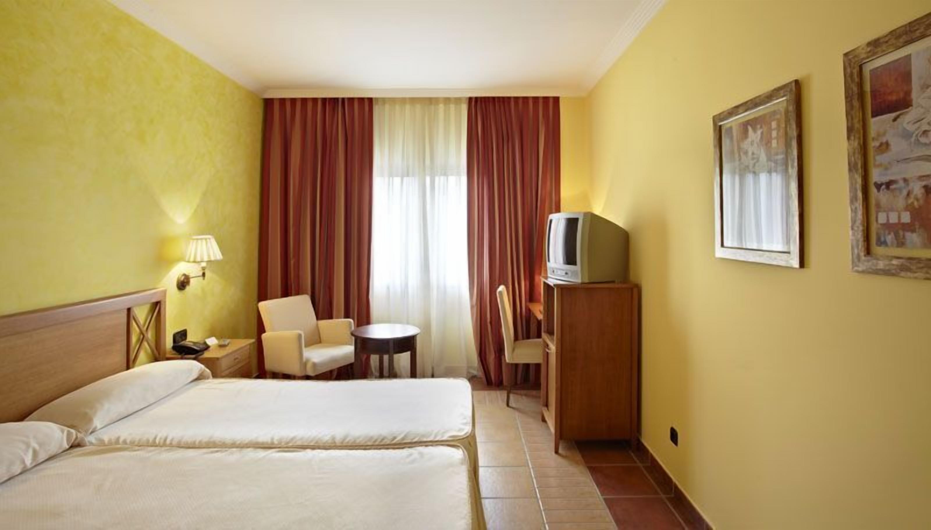 A hotel room with two beds, a desk with a television, a chair, a small table, and a lamp.