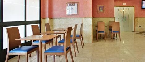 Un comedor con mesas y sillas de madera, suelo de baldosas y pared rosa decorada con marcos.