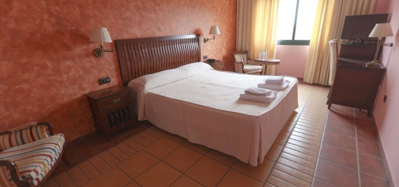 A hotel room with a large bed, a wooden headboard, two bedside tables with lamps, a small chair, a desk, and a TV.
