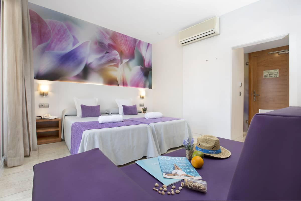 Una habitación con dos camas, un amplio mural floral abstracto, una puerta de madera y una pequeña mesa con un sombrero y un libro.