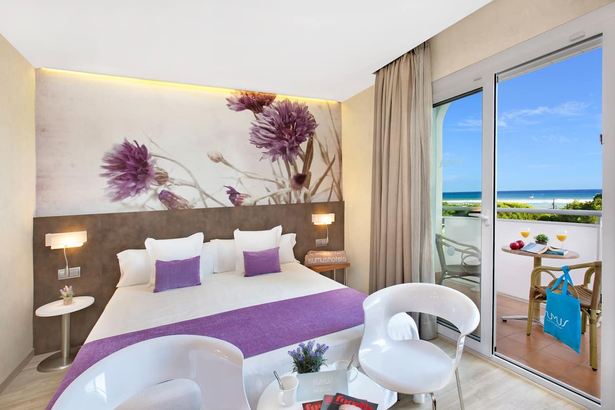 Una habitación de hotel moderna con una cama grande, un ventanal que da a un balcón con vistas al mar y una mesita con una bebida y un bolso.