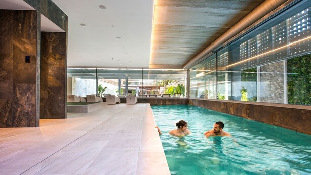Une piscine intérieure à l’eau claire, dotée d’une architecture moderne et de grandes fenêtres offrant une vue sur la verdure et les bâtiments environnants.