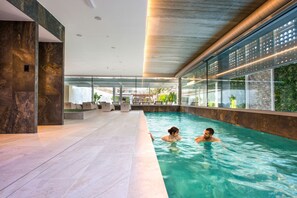 An indoor swimming pool with clear water, modern architecture, and large windows offering views of greenery and buildings outside. - Sumus Hotel Monteplaya & Spa 4S - Adults Only (Malgrat de Mar)