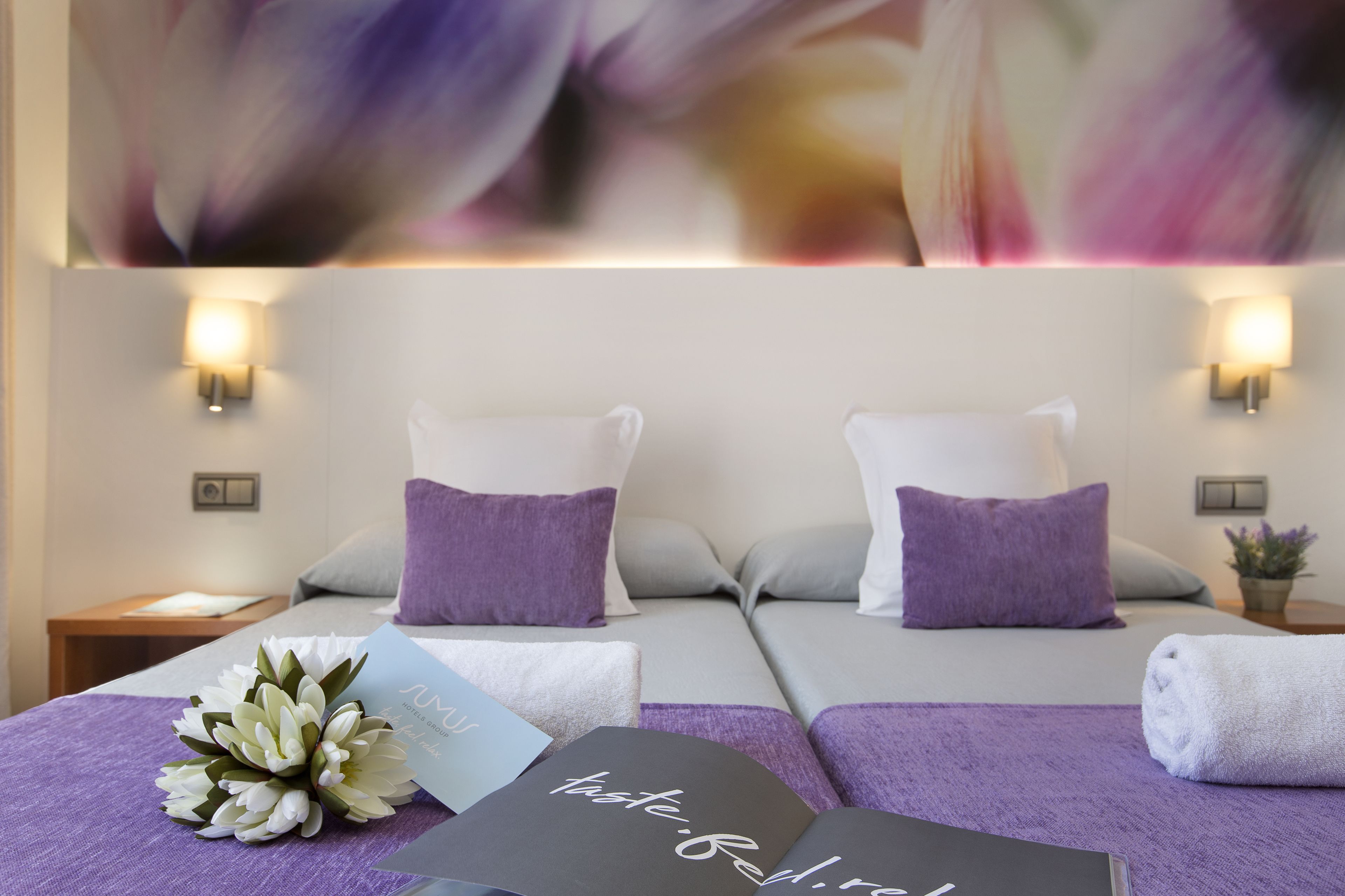 Una habitación de hotel con dos camas, cada una con un cojín morado, una mesita de noche con una lámpara y un ramo de flores sobre la cama.
