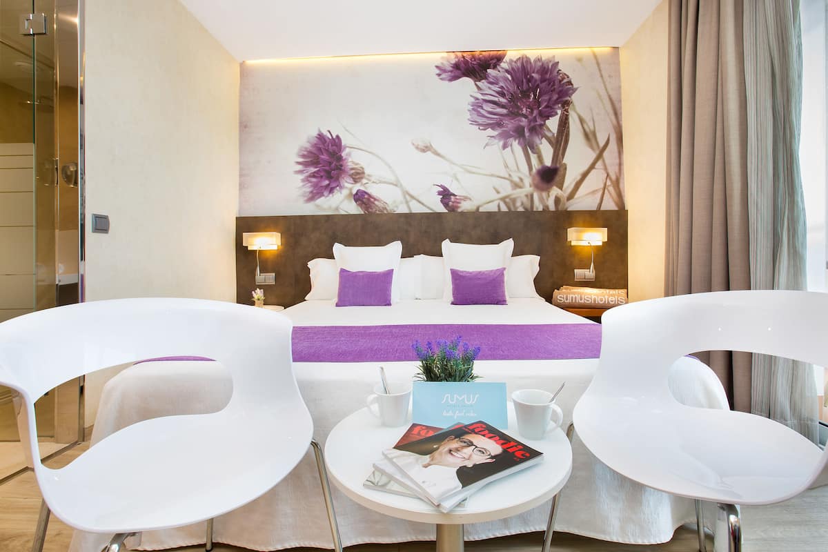Una habitación de hotel moderna con una cama grande, dos sillas blancas, una mesita con una revista y una pared decorada con flores púrpuras.