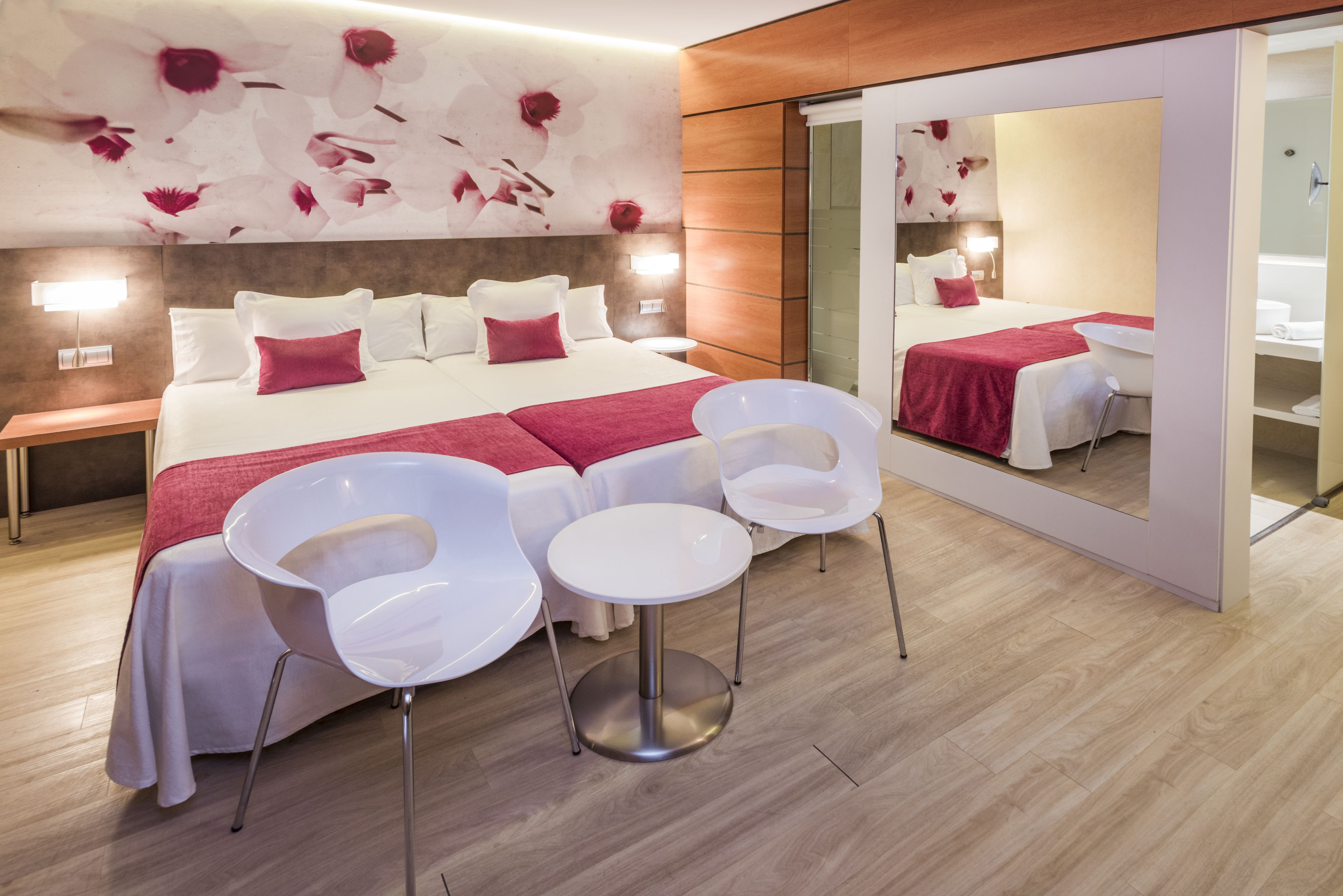 Una habitación de hotel moderna con una cama grande, sillas blancas, una mesita pequeña y un espejo amplio.