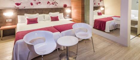Una habitación de hotel moderna con una cama grande, sillas blancas, una mesita pequeña y un espejo amplio.