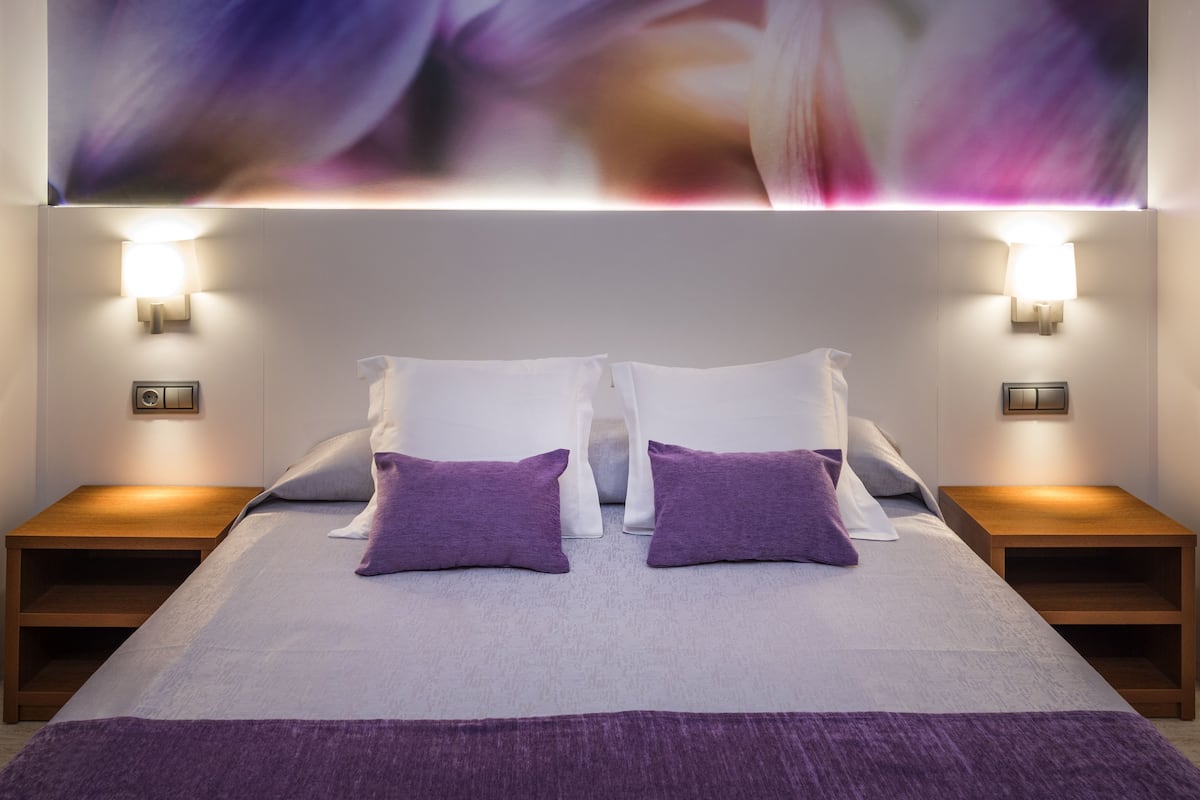 Una cama bien hecha con almohadas blancas y moradas, dos mesitas de noche con lámparas y un cuadro abstracto grande sobre la cama.