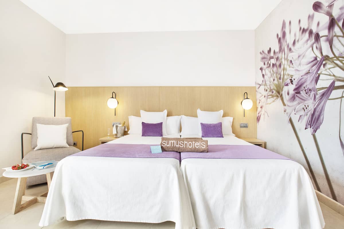 Habitación de hotel con dos camas, una mesita con un plato de fresas, una silla y una pared con un mural floral.