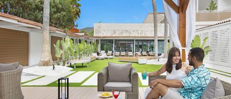 A covered outdoor seating area with wicker furniture, a table with food and drinks, and a view of a modern building in the background.