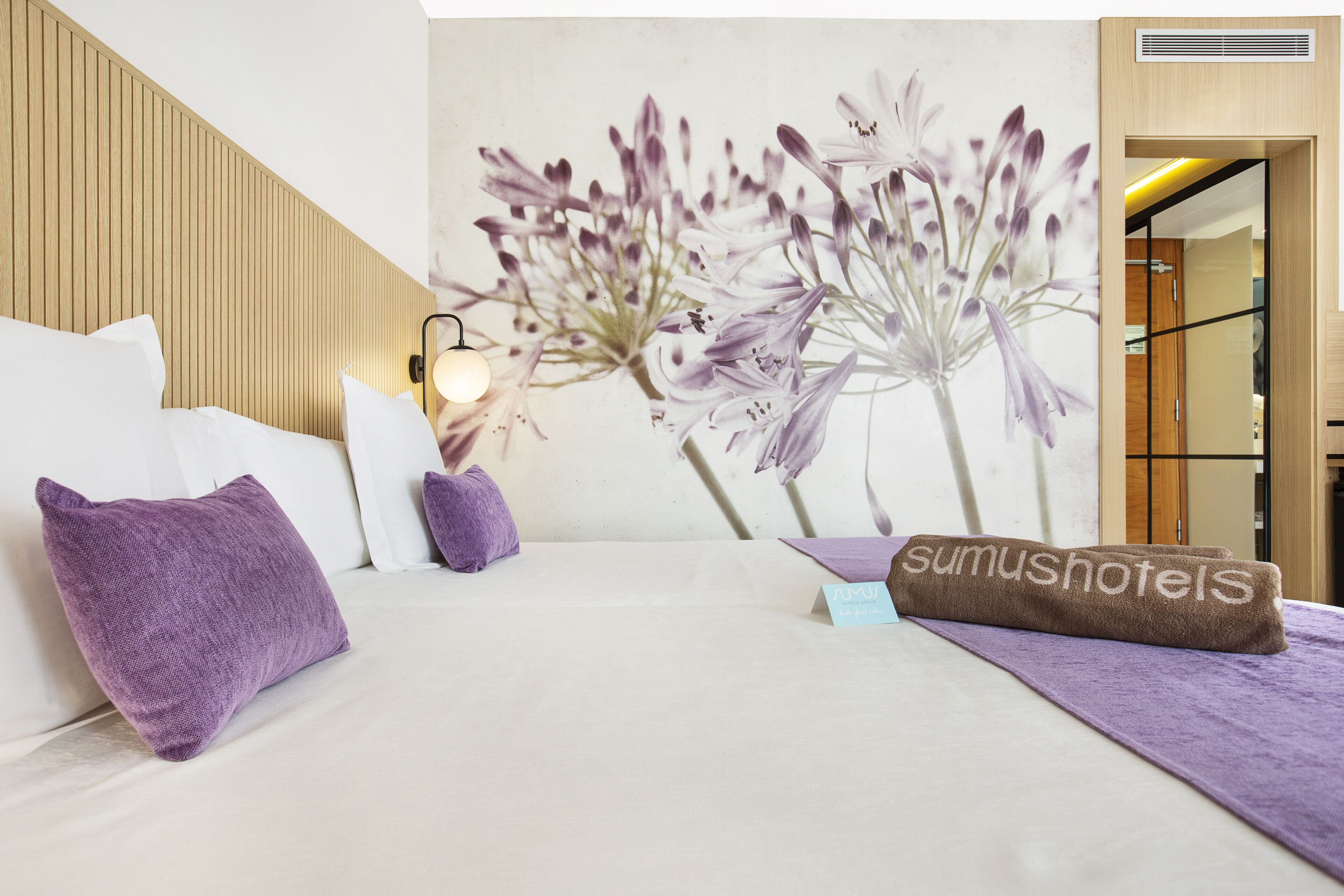 Ein Hotelzimmer mit einem Bett, Kissen, einer Wandlampe und einem floralen Wandgemälde.