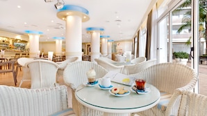 Un restaurant avec des chaises en osier et une table en verre, un bar à l’arrière-plan et de grandes fenêtres donnant sur des palmiers.