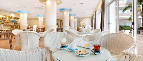 Un restaurante con sillas de mimbre y una mesa de vidrio, un bar al fondo y ventanas amplias con vistas a palmeras.