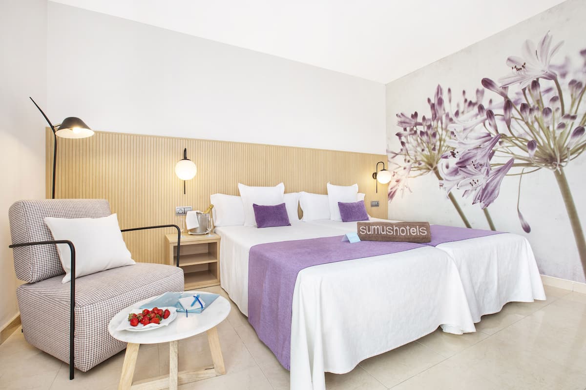 Una habitación de hotel con una cama, dos mesitas de noche, una silla, una pequeña mesa con frutero y una lámpara de pared.