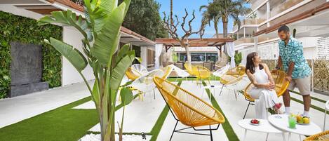 A modern outdoor seating area with yellow chairs, white tables, and a small garden with artificial grass and a palm tree.