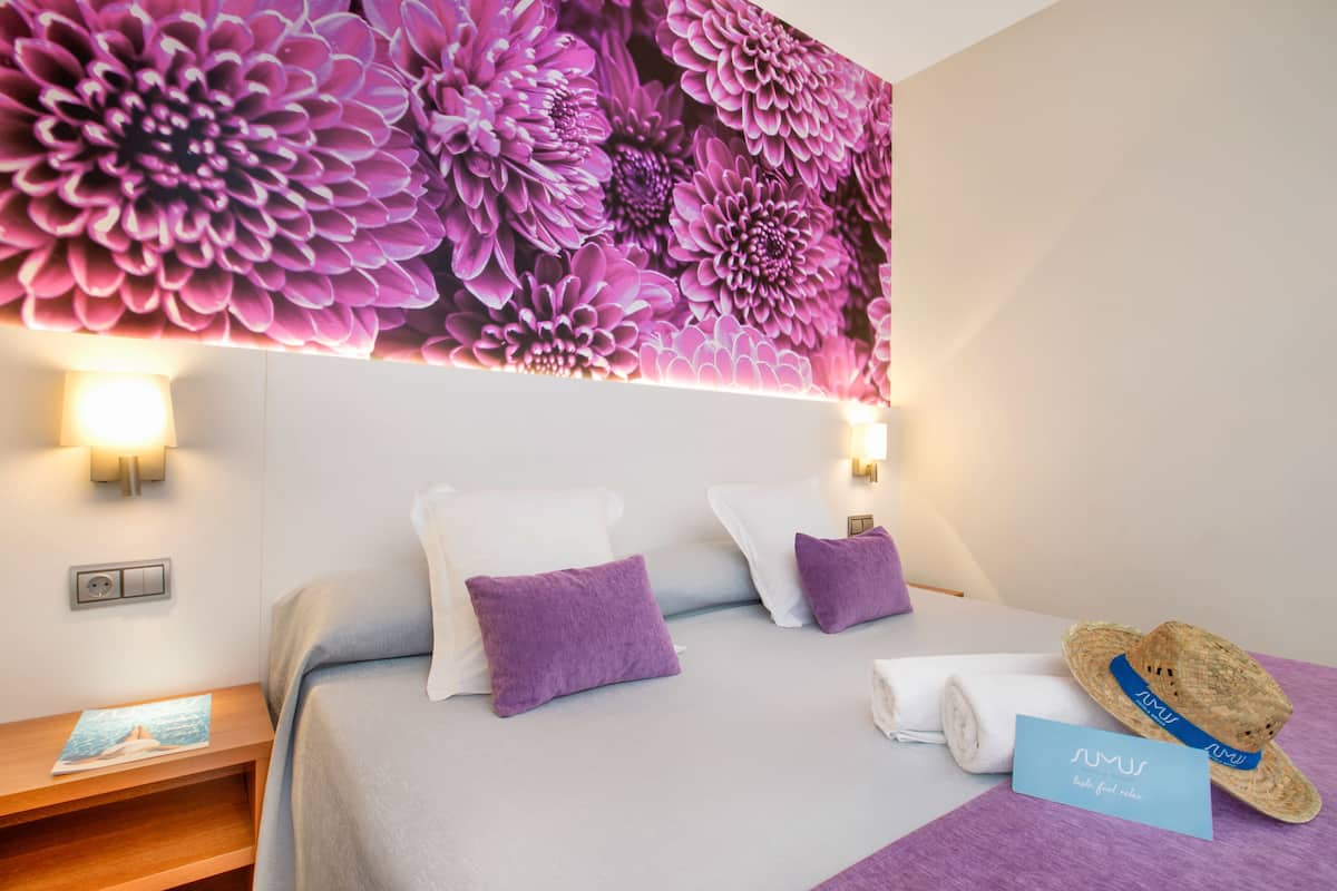 Habitación de hotel con un gran mural floral, una cama con ropa de cama blanca y morada, una mesita de noche de madera, un sombrero y una toalla.