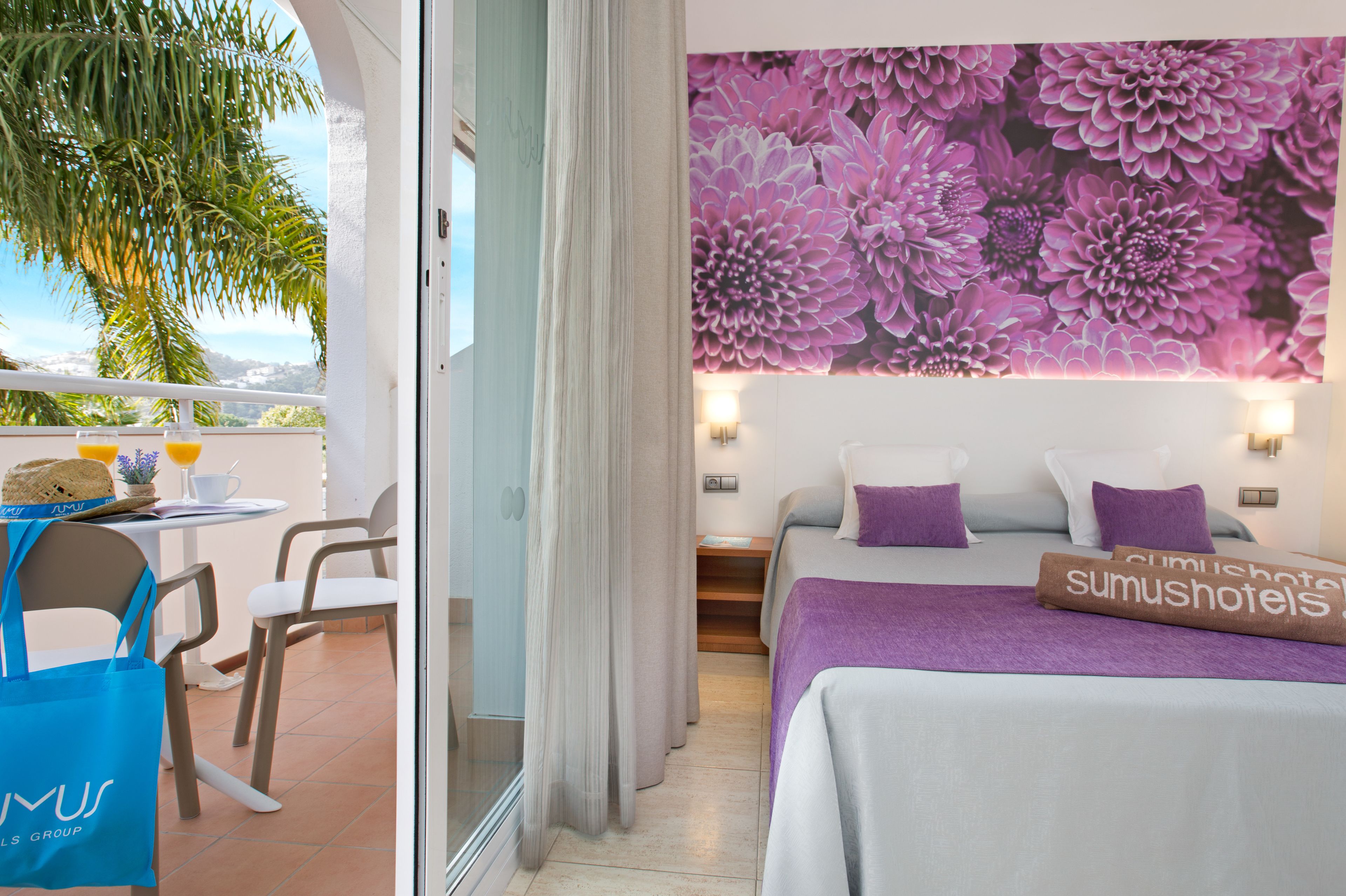 Habitación de hotel con balcón, cama con ropa de cama morada y un mural floral en la pared.