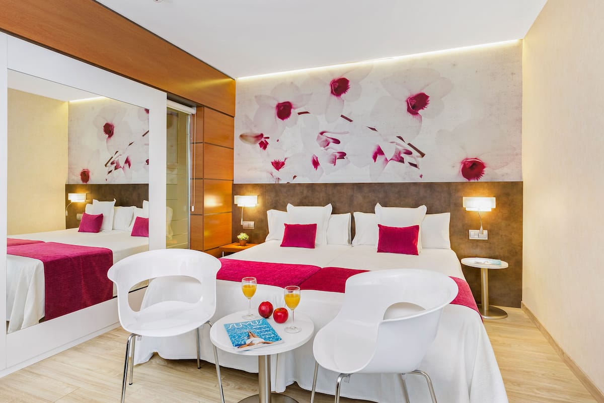 Habitación de hotel con una cama grande, dos sillas blancas, una mesita con bebidas y manzanas, y una pared con diseño floral.