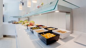 A modern buffet setup with various food items in trays, a glass railing, and pendant lighting. - Beverly Park Hotel & Spa (Blanes)