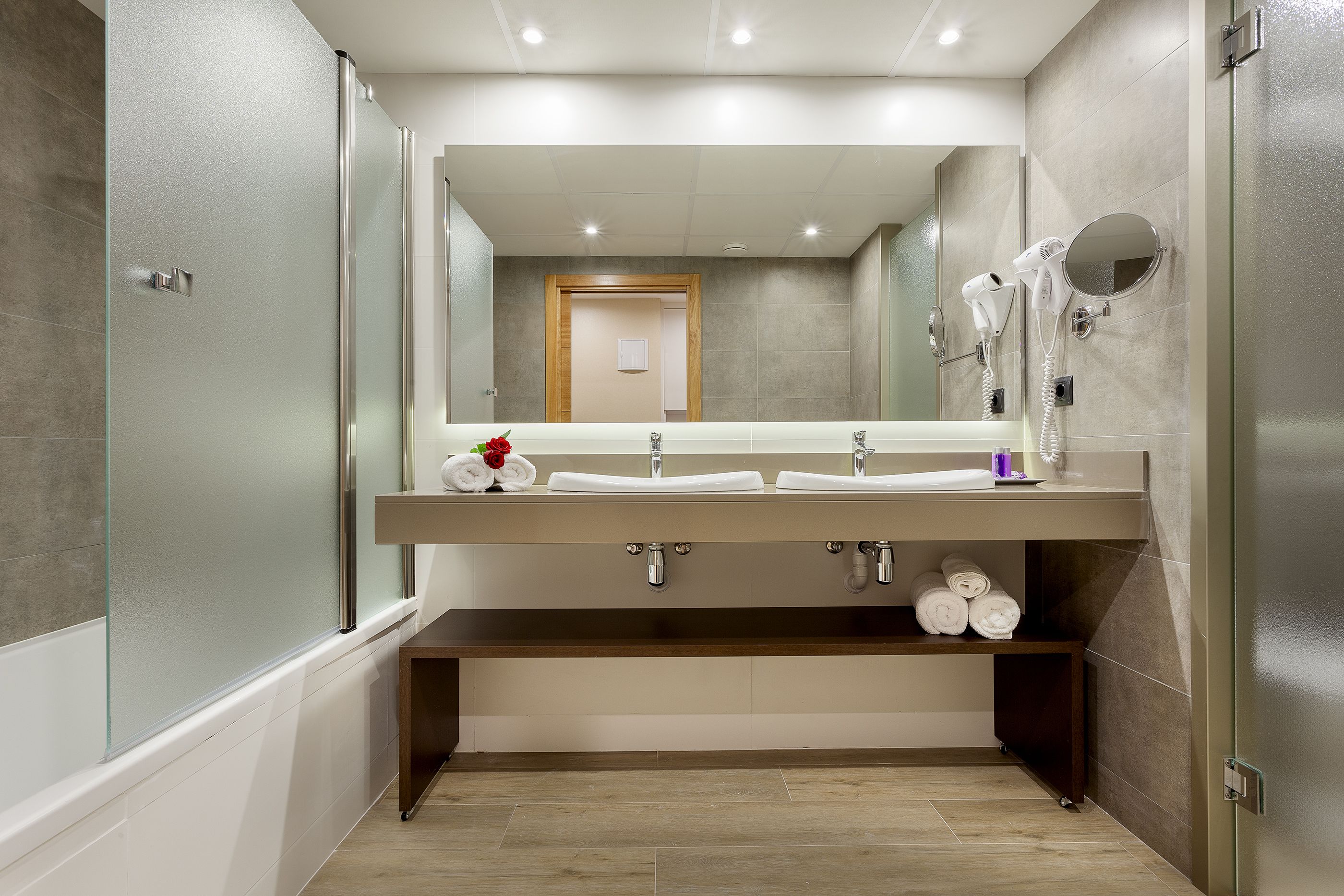 A modern bathroom with a large mirror, two sinks, a towel rack, and a shower area.