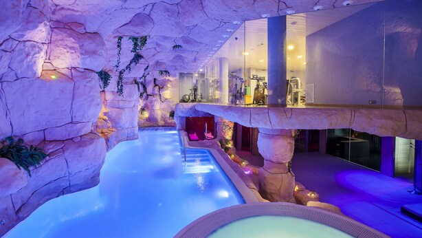 An indoor pool with a rocky cave-like ceiling, clear water, and a glass railing overlooking the pool. - Beverly Park Hotel & Spa (Blanes)
