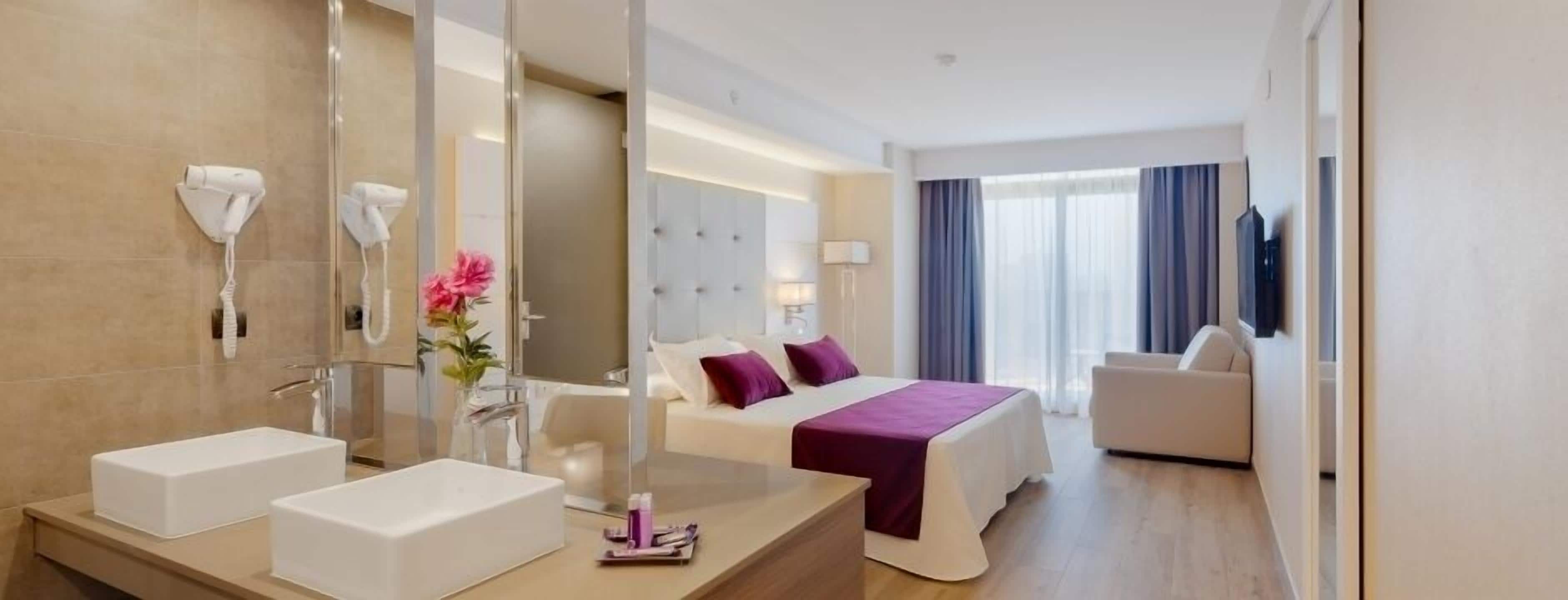 Een hotelkamer met een grote spiegel, twee wastafels, een bed met paars beddengoed, een nachtkastje, een lamp en een uitzicht op de stad door grote ramen.