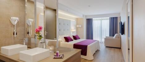 A hotel room with a large mirror, two sinks, a bed with purple bedding, a bedside table, a lamp, and a view of the city through large windows.