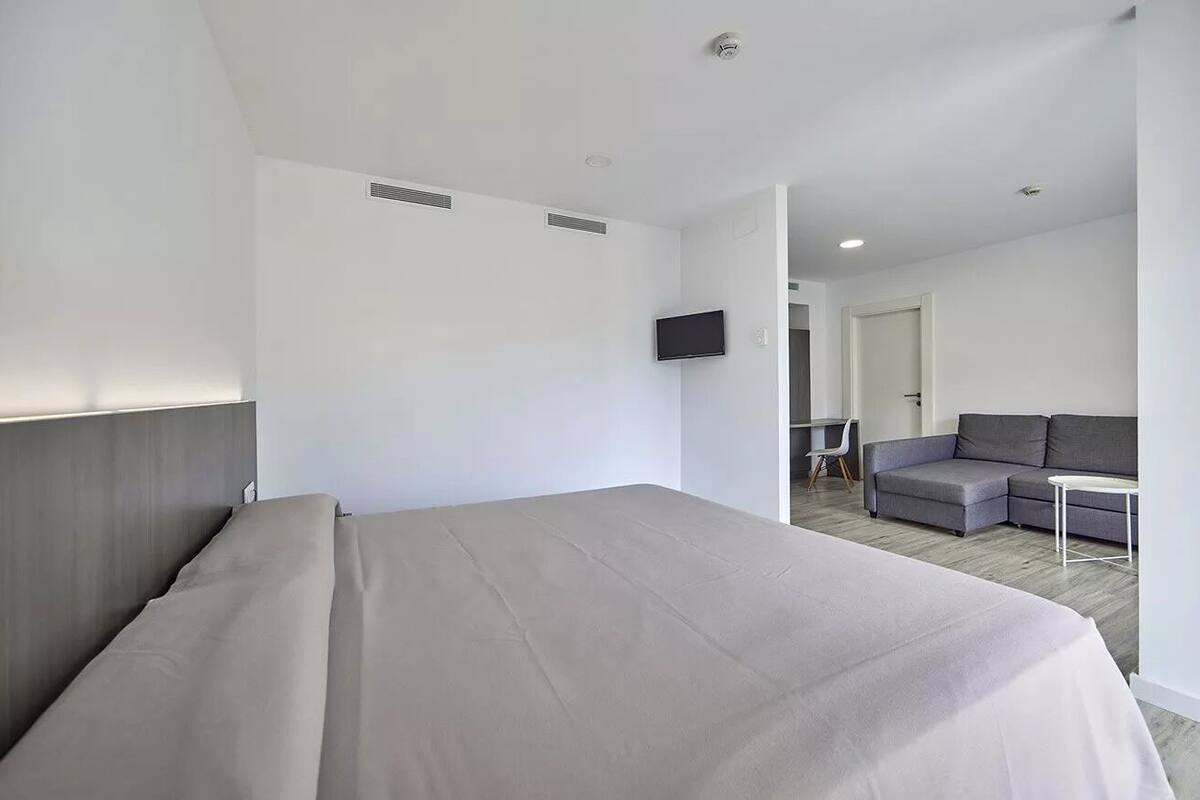 Une chambre d’hôtel avec un lit, un canapé gris, une petite table et un téléviseur fixé au mur.