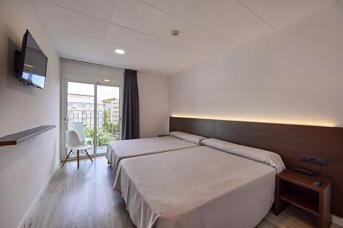 Une chambre d’hôtel avec deux lits, un téléviseur fixé au mur, une chaise et une vue sur l’extérieur.