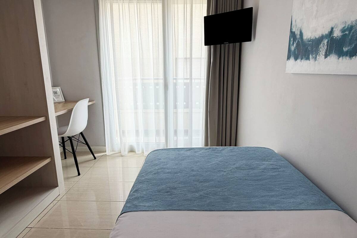 Un dormitorio con cama, escritorio, silla, televisor y ventana con cortinas.