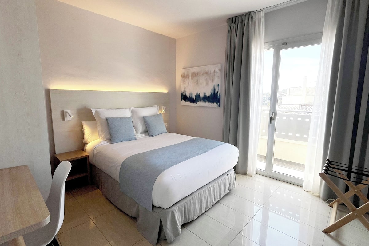 Habitación de hotel con cama, escritorio, silla, vistas al exterior y un cuadro en la pared.