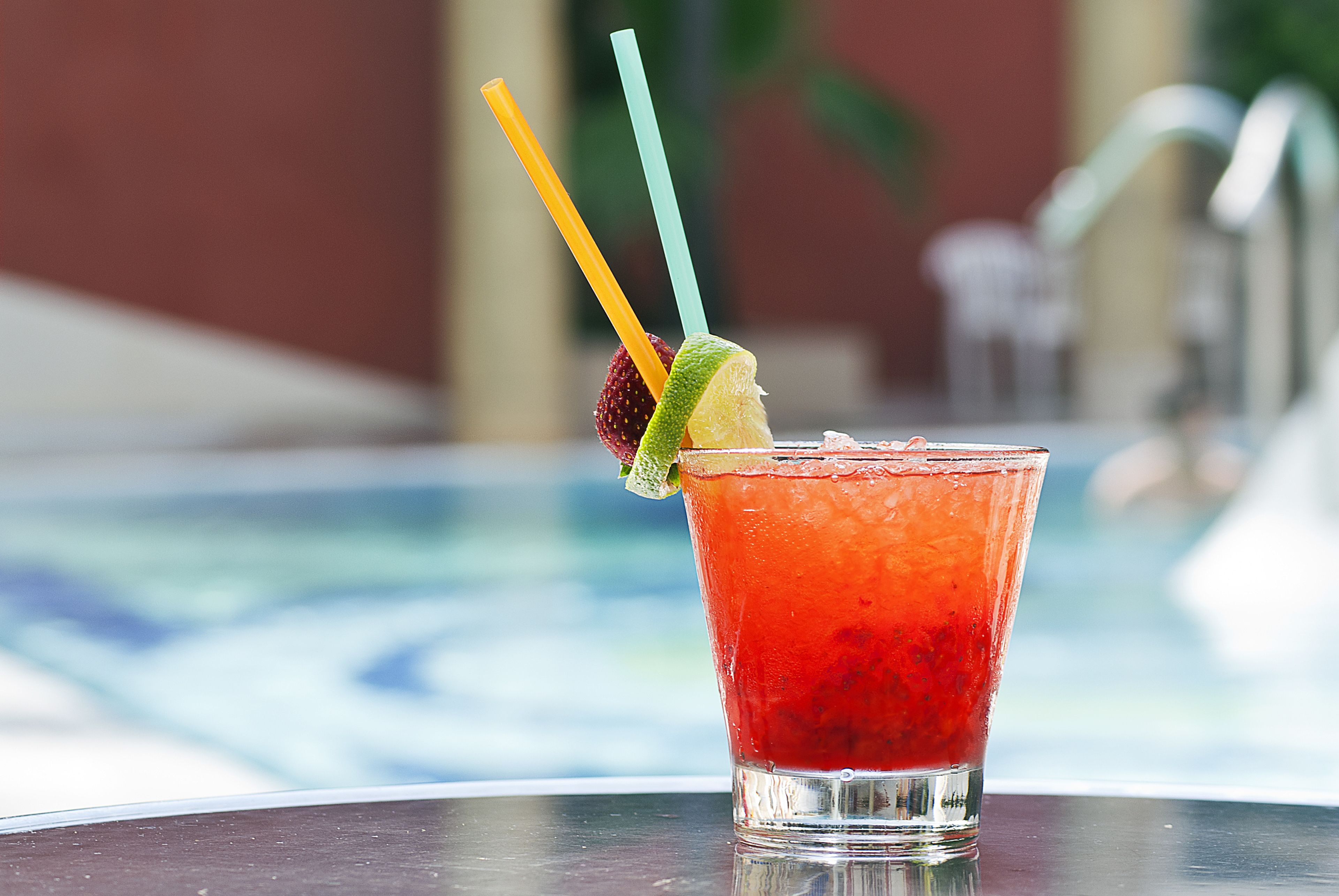 A cocktail with a lime wedge and a strawberry, served in a glass with two straws, placed on a table near a pool.