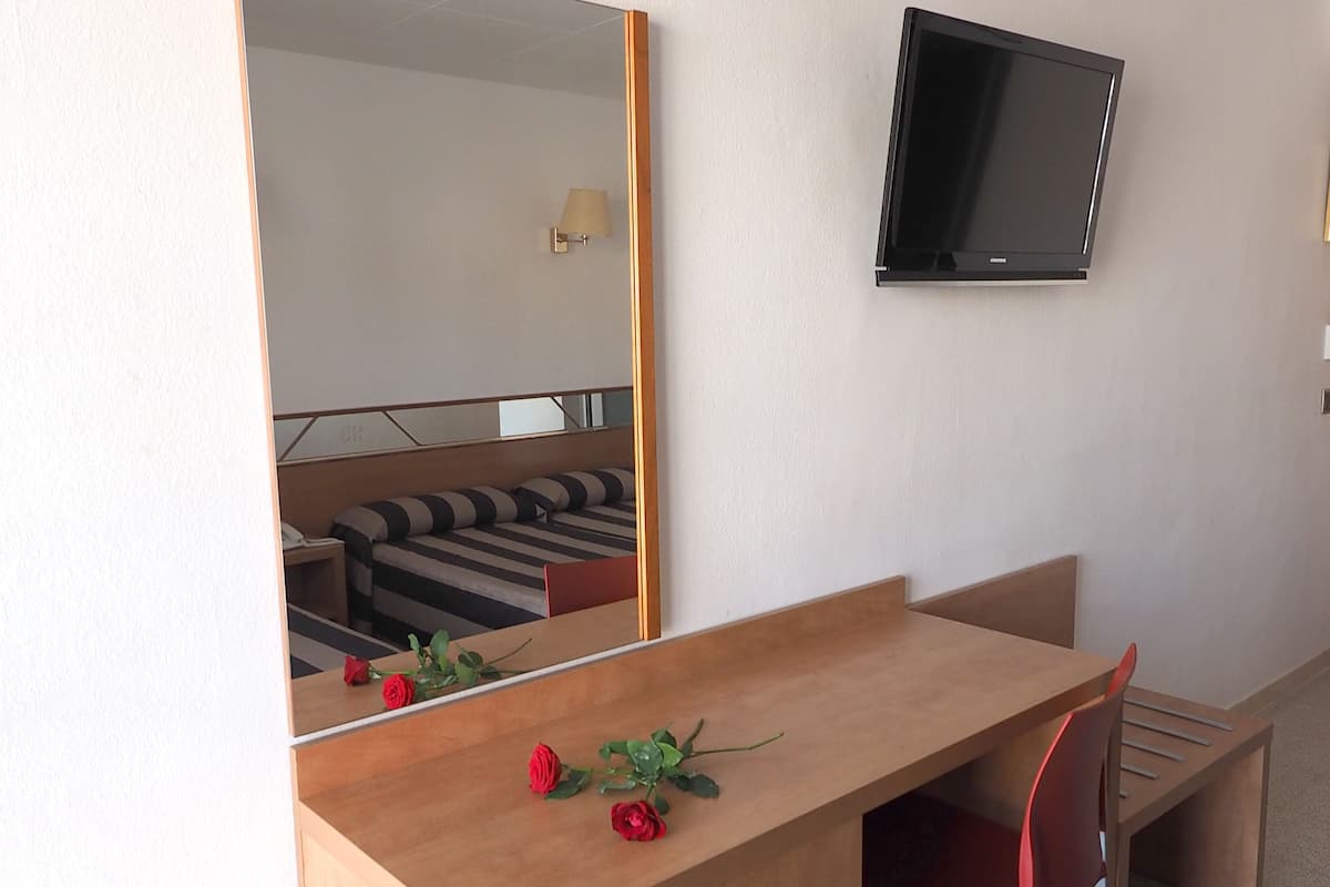 A modern hotel room with a wooden desk, a red chair, a television mounted on the wall, and a large mirror reflecting the room.