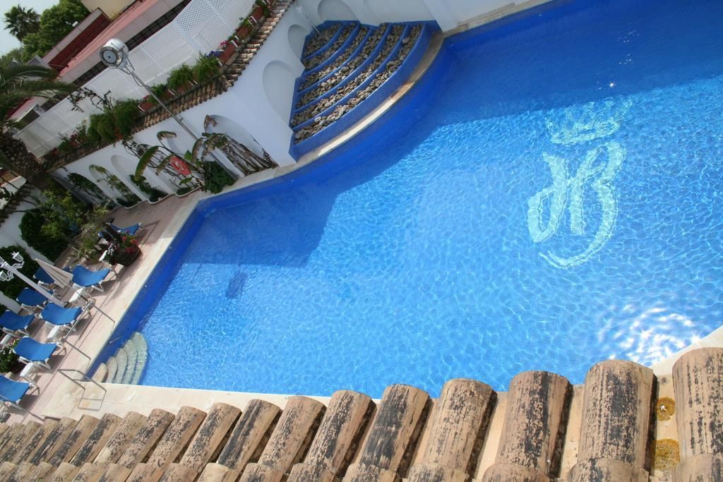 A swimming pool with a clear blue water surface, surrounded by lounge chairs and a white wall with a decorative pattern.