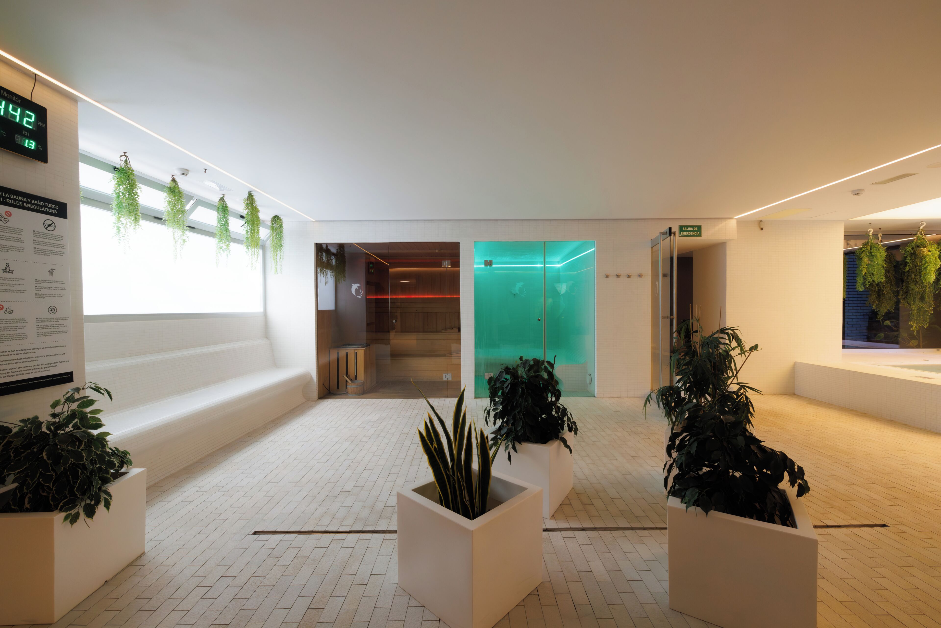 Un vestíbulo moderno con un reloj digital, plantas en macetas y una puerta de vidrio que da a una habitación.