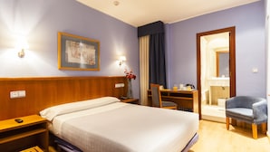 A hotel room with a large bed, a desk with a chair, a mirror, and a bathroom visible through an open door. - City Express Covadonga (Oviedo)