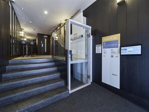 Entrada de un hotel con puerta de vidrio, una estación de recepción y una escalera que conduce al piso superior.