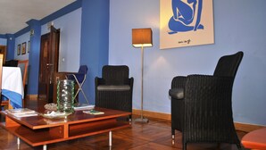 A room with blue walls, wicker chairs, a wooden coffee table, a floor lamp, and a framed artwork. - City Express Covadonga (Oviedo)