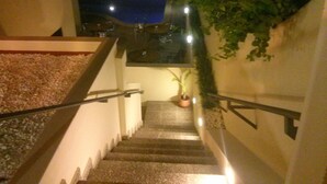 Una escalera con una planta en maceta al pie, barandilla a la derecha y una fuente de luz en lo alto.