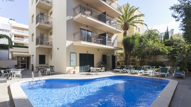 Una piscina con sillas de descanso y mesas, rodeada por un edificio con balcones y una palmera.
