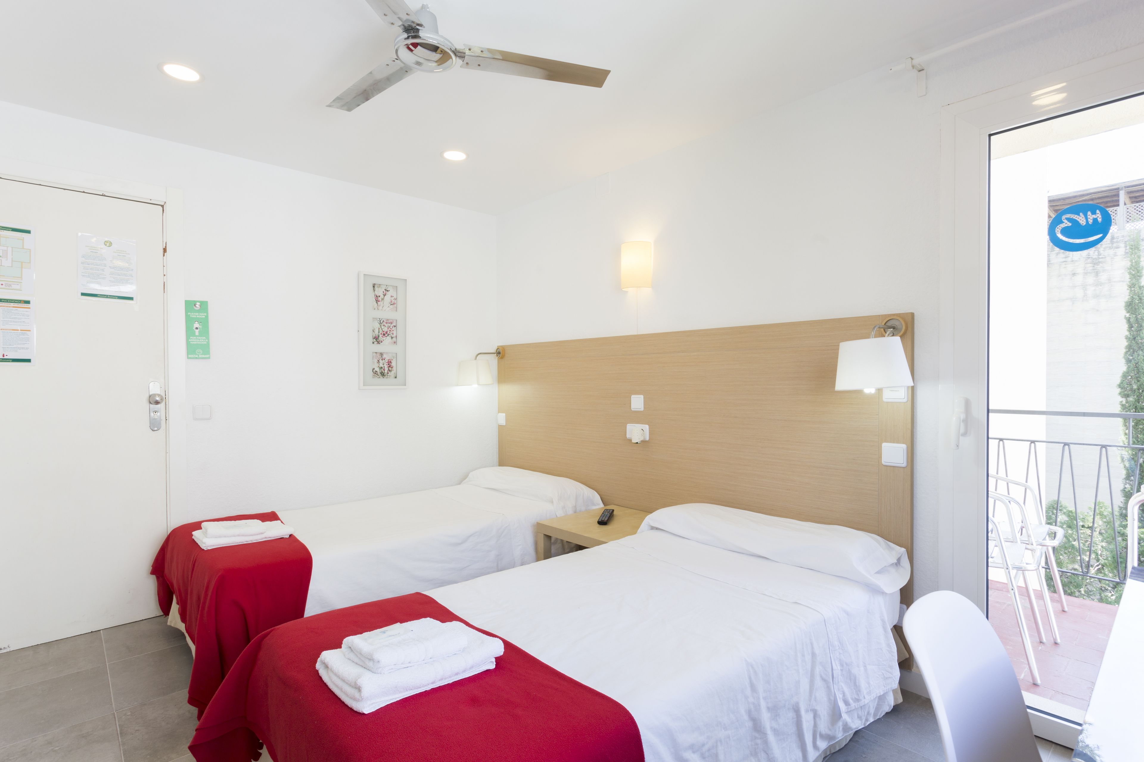 Habitación de hotel con dos camas, ventilador de techo, balcón con sillas y vistas al exterior.