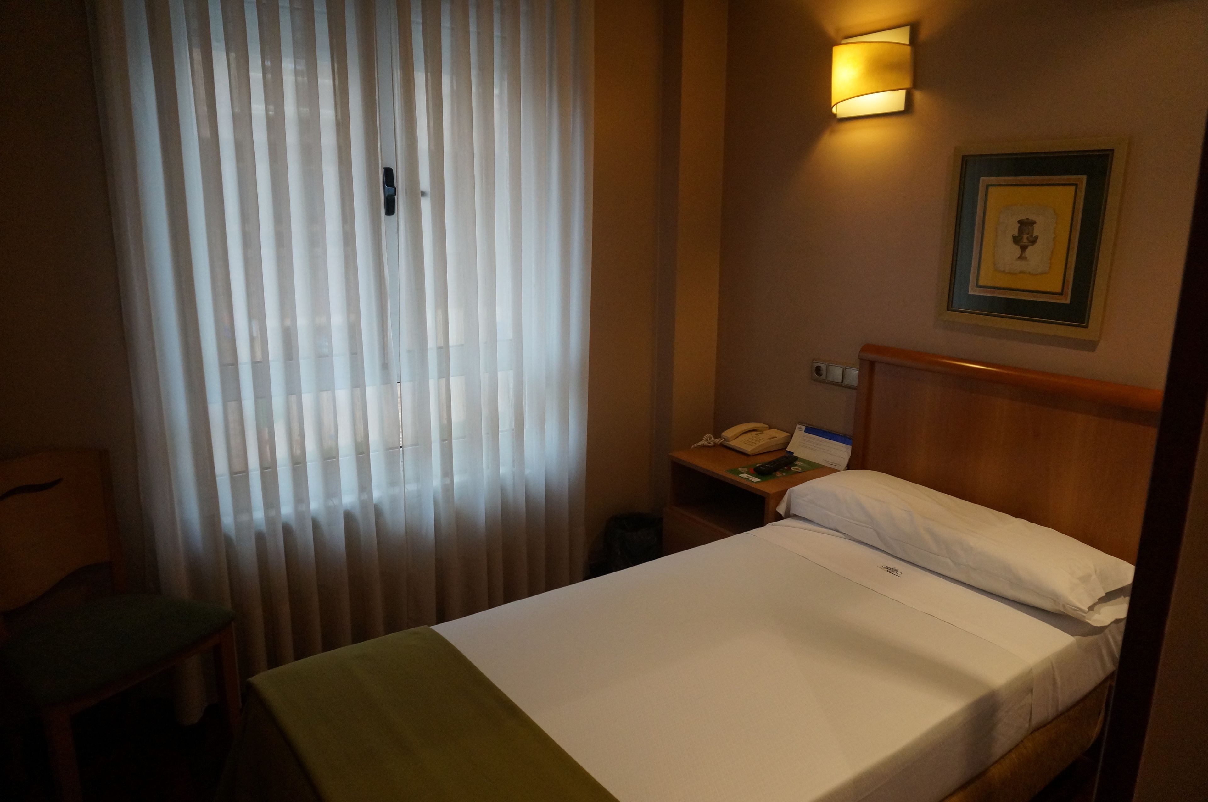 Habitación de hotel con cama, mesita de noche con lámpara, un cuadro enmarcado en la pared y una ventana con cortinas.