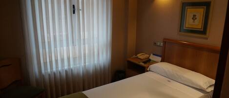 Habitación de hotel con cama, mesita de noche con lámpara, un cuadro enmarcado en la pared y una ventana con cortinas.