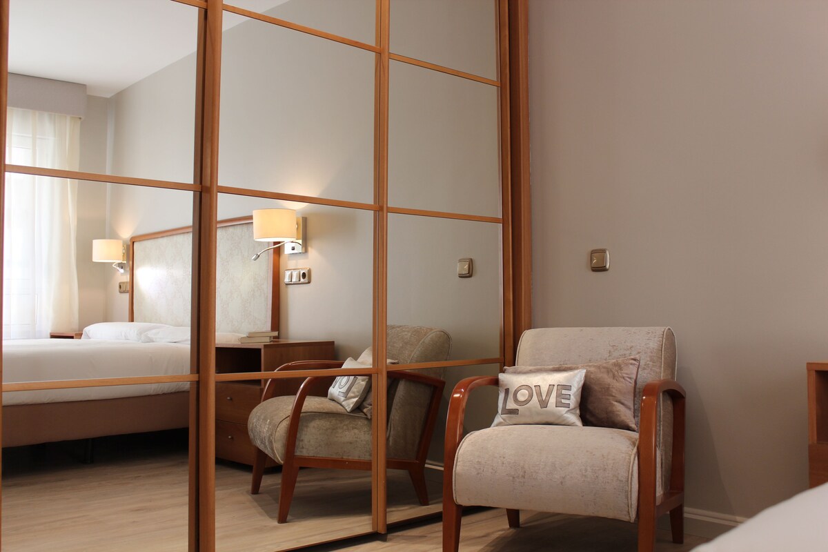 A hotel room with a bed, a chair with a pillow, a nightstand, and a wall-mounted lamp.