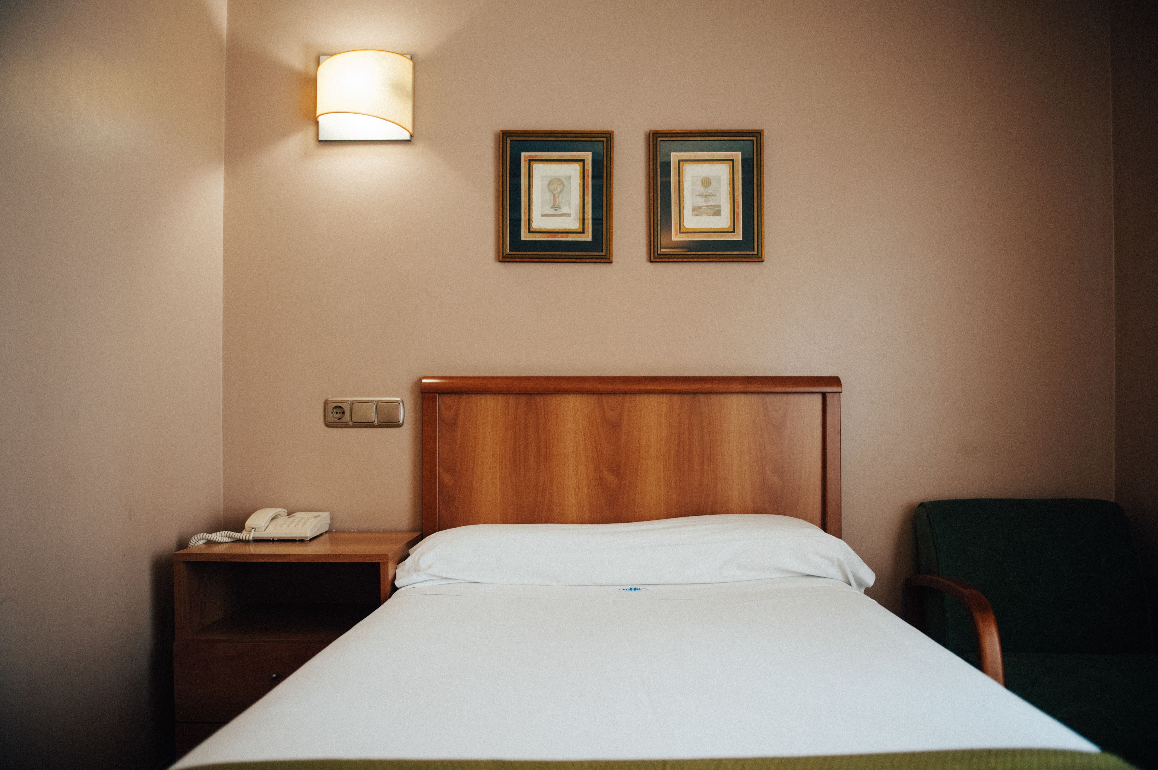 Habitació d'hotel amb un capçal de llit de fusta, una taula de nit amb un telèfon, una cadira verda i dos quadres al mur.