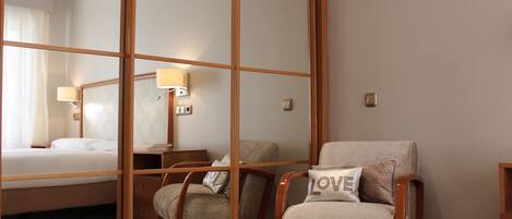 A hotel room with a bed, a chair with a pillow, a nightstand, and a wall-mounted lamp.