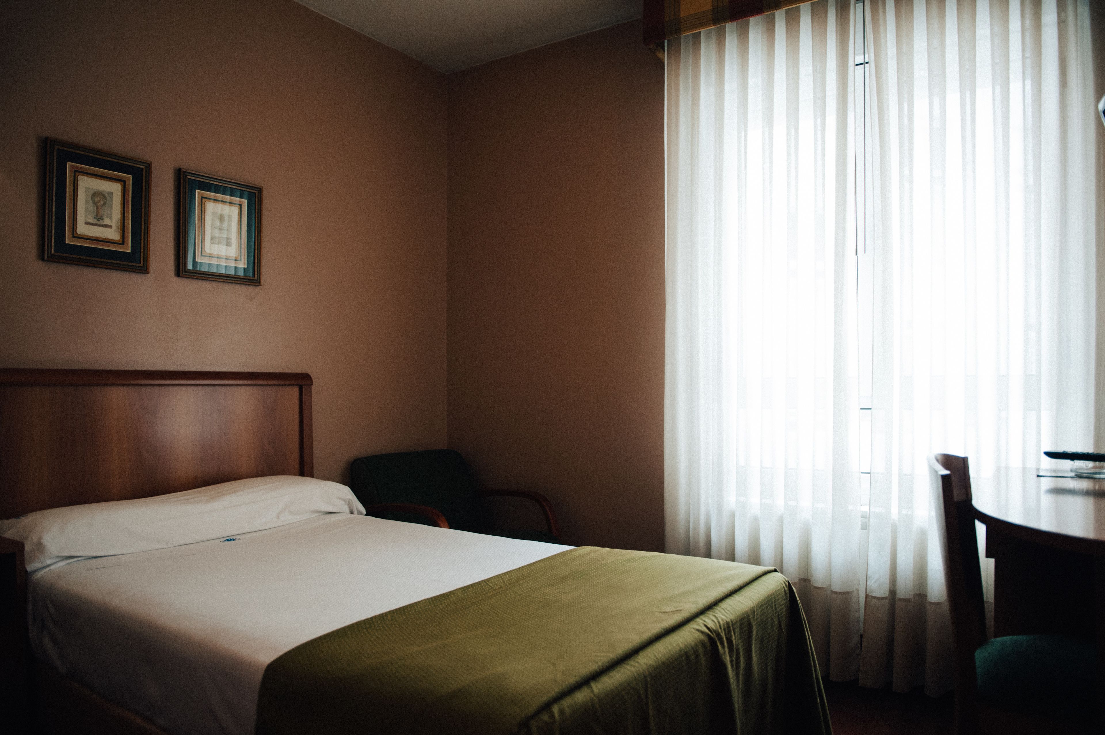 A hotel room with a bed, two framed pictures on the wall, a chair, a table, and a window with sheer curtains.
