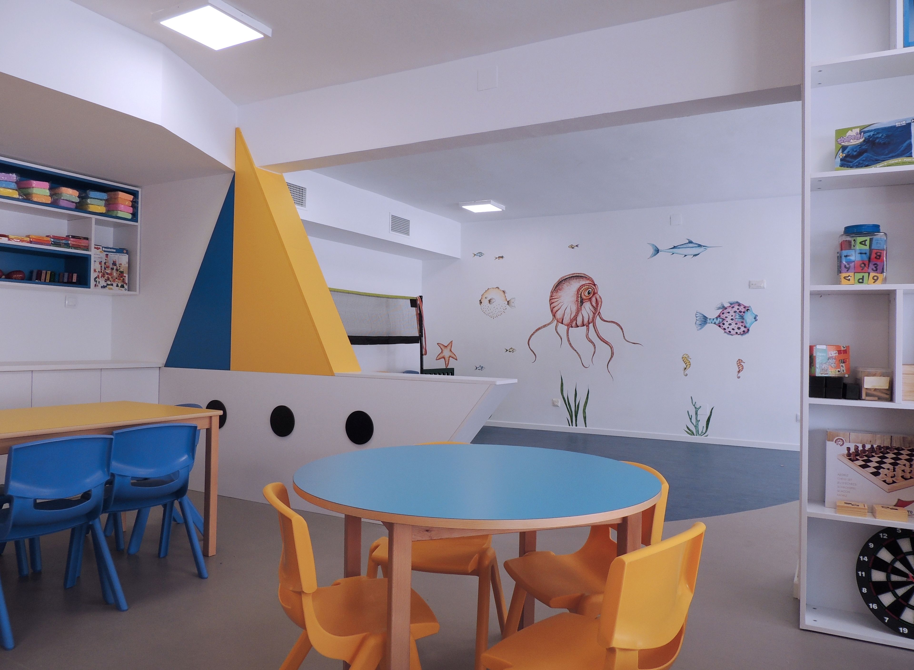 Un cuarto de juegos infantil con una mesa azul, sillas amarillas y una estantería llena de juguetes.